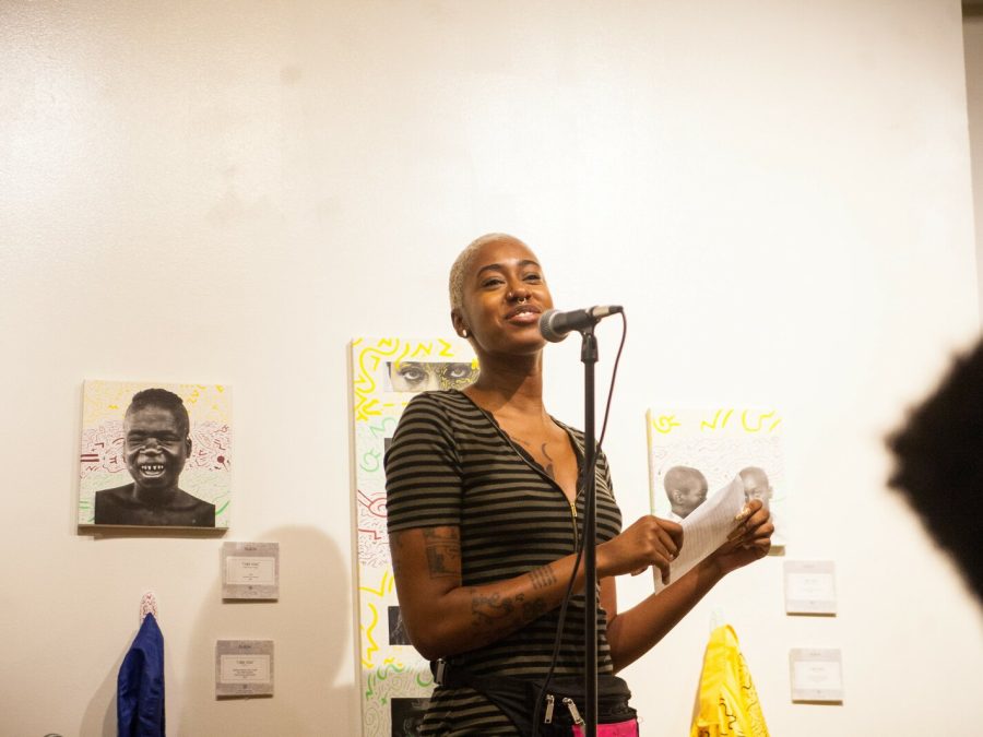 A brown-skinned person with short blond hair stands in front of a microphone with papers and images taped to a white wall behind them.