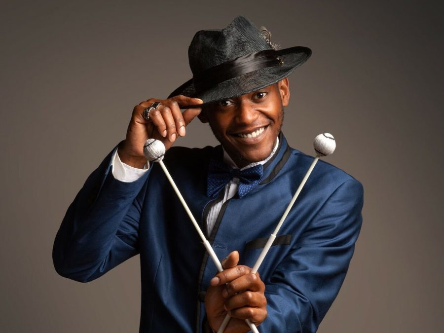A Black man holds a set of mallets in his left hand towards the camera while tipping his gray hat with his right hand.