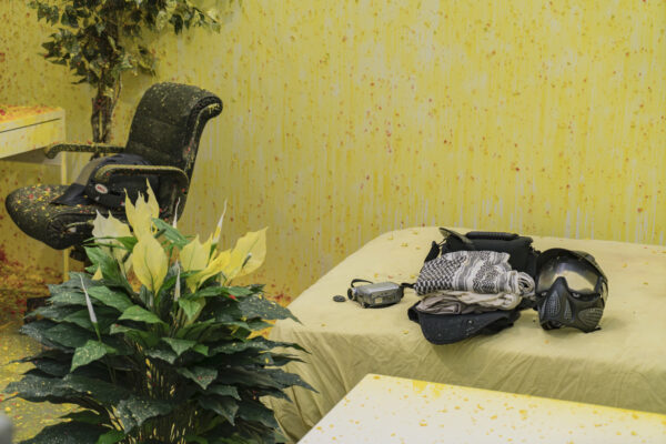 A yellow paint splattered room with flowers in the foreground and a bed and desk chair in the background.