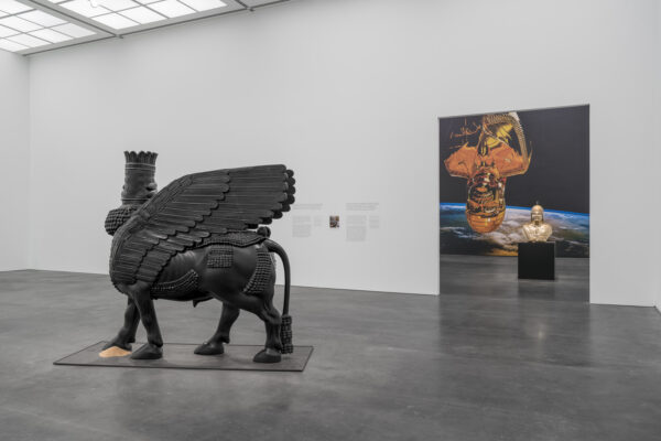 Installation view of two rooms of a gallery the first room contains a black lamassu and the room behind it contains two artworks of Saddam Hussein