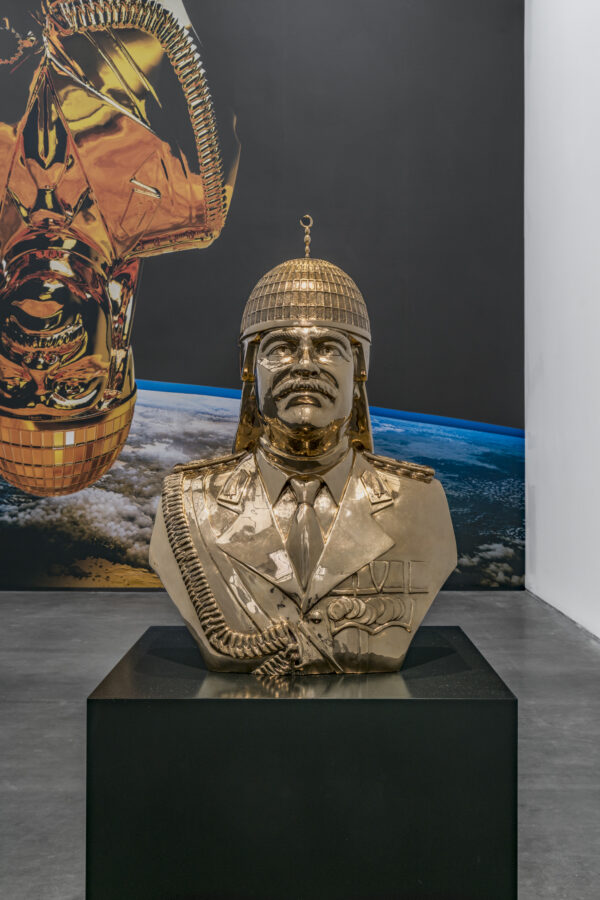 A golden bust of Saddam Hussein on a black pedestal. Behind it appears an image of that bust upside down in outer space