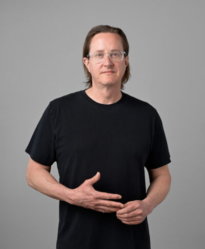 A portrait of a light-skinned man in a black shirt and jeans with slicked back hair posing against a gray wall