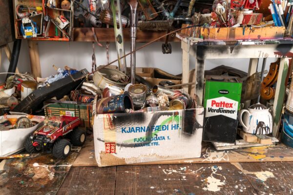 A cluttered workspace filled with various tools and objects. A toy truck is visible on the floor. A cardboard box labeled 
