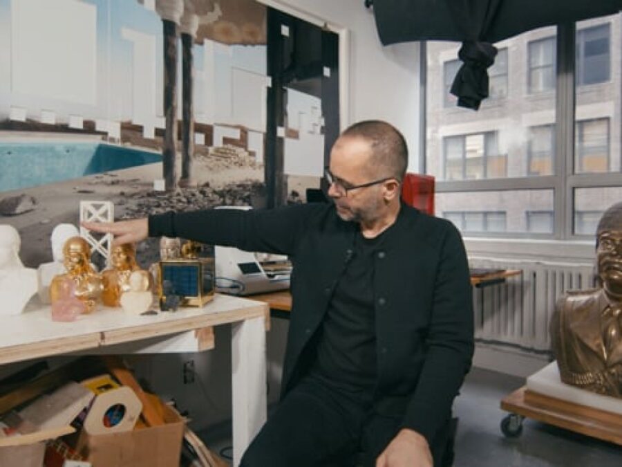 A man with shorthair and glasses dressed in all black in a studio filled with busts of Saddam Hussein