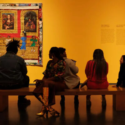 A group of people sitting on a bench in an orange room looking at a colorful quilt.