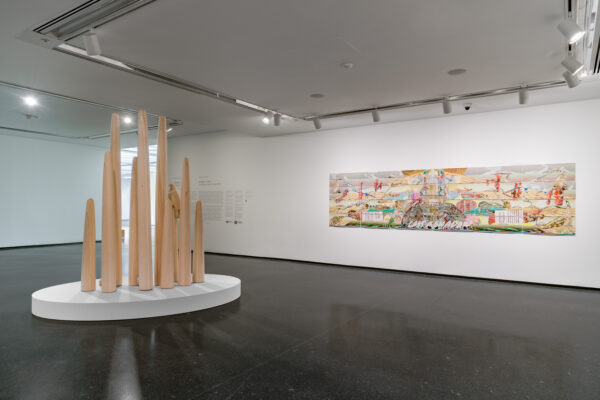 A series of various sized wooden spires on a white platform in the middle of a gallery space near a wide artwork hanging on the wall