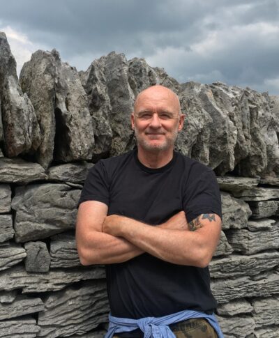 A person poses with their arms crossed next to a wall of rocks