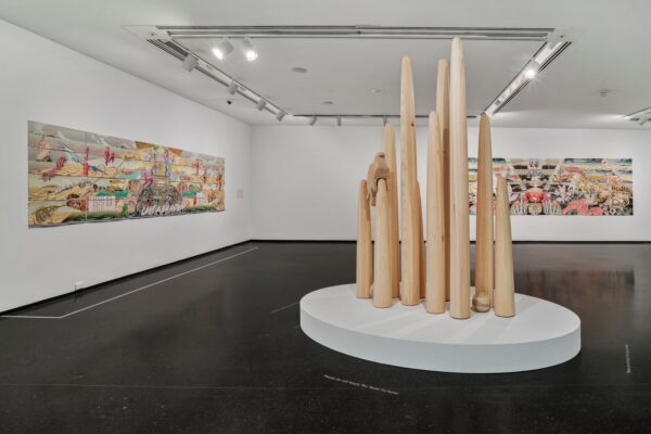 A series of various sized wooden spires with a carved bird sitting on one of them on a white platform in the middle of a gallery space with two paintings hanging on two adjacent walls