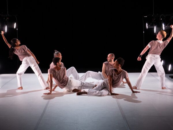 Six performers in matching outfits on a stage. Four sit in a circle in the middle while two others stand on either side with one arm stretched out giving the illusion that they are forming a W together.