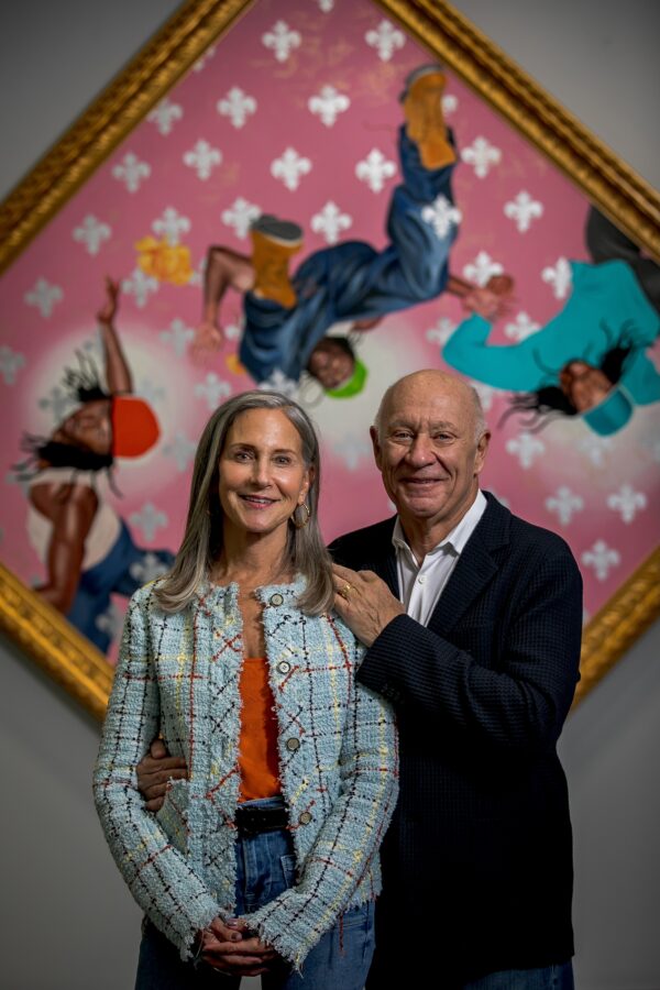 A man and a woman stand in front of a diamond shaped painting. The main has his arms on the shoulder of the woman.
