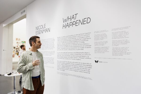 A person holding a drink stands in front of exhibition introduction text on a wall titled What Happened