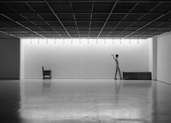 A black and white image of a nude person walking between two objects in an otherwise empty gallery space