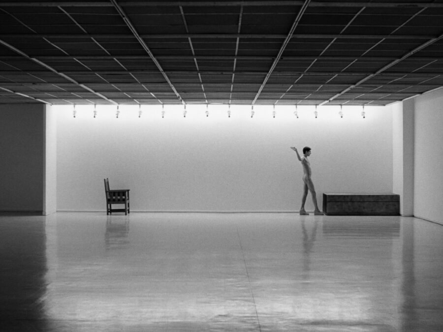 A black and white image of a nude person walking between two objects in an otherwise empty gallery space
