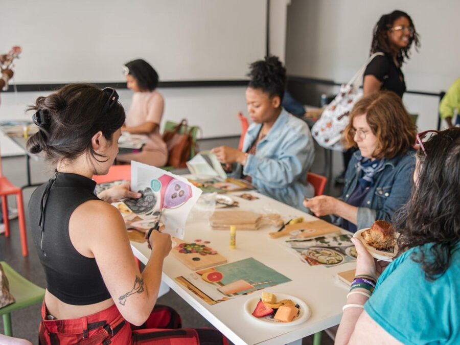 A group of people sit at a table with magazine pages scattered across it. Some are working on cutting out images from these pages.