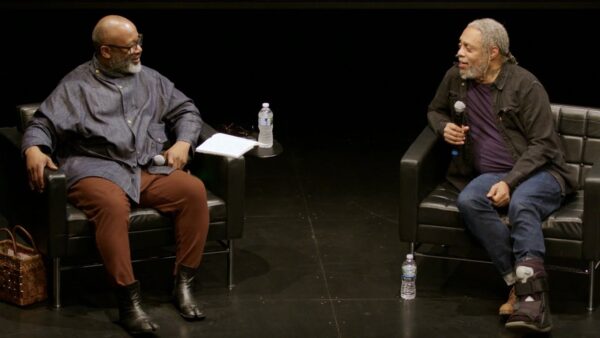 Two Black men sit on an all-black stage conversing. Each holds a microphone.