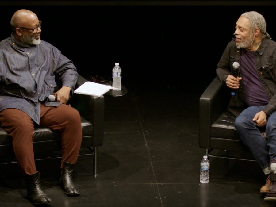 Two Black men sit on an all-black stage conversing. Each holds a microphone.
