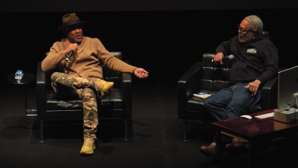 Two Black men sit on an all-black stage conversing. Each holds a microphone.