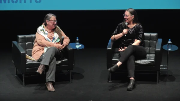 Two people sit on black chairs on a black stage holding microphones