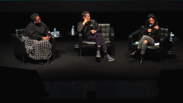 Three people of various ethnicities sit in black chairs on an all-black stage holding microphones