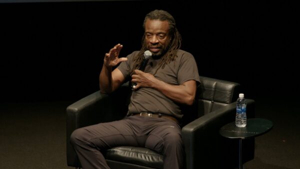 A Black man wearing all black sits on an all-black stage. He raises his right arm while holding a microphone with his left hand