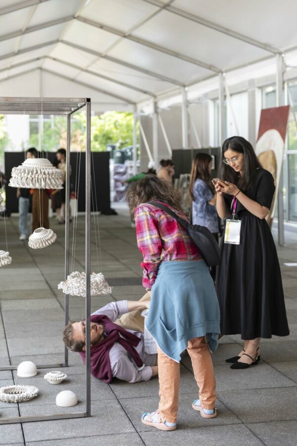 White sculptures hang from a metal structure under a white canopy. Two people gather around the artworks and a man lying on the ground looking at one of them.