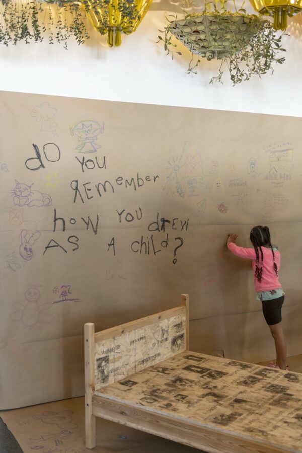 A person writes on a brown wall filled with other paper and a wooden bed filled with black and white images and text. The largest text written on the wall reads do you remember how you drew as a child