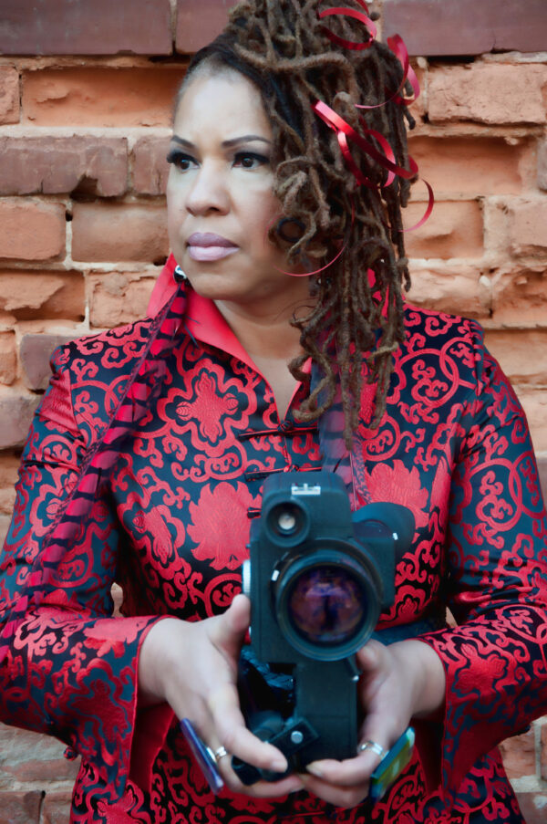 A brown-skinned, female presenting person in a bright red shirt looks off to the side while holding a camera