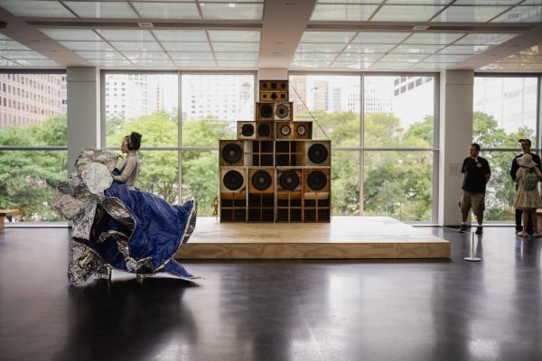 A person in a blue and silver metallic dress walks past an arrangement of old speakers