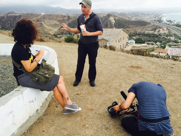Three people on a sandy hill overlooking a building and a dusty road. One is sitting on a stone bench looking at a person standing while being filmed by someone sitting on the ground with a camera