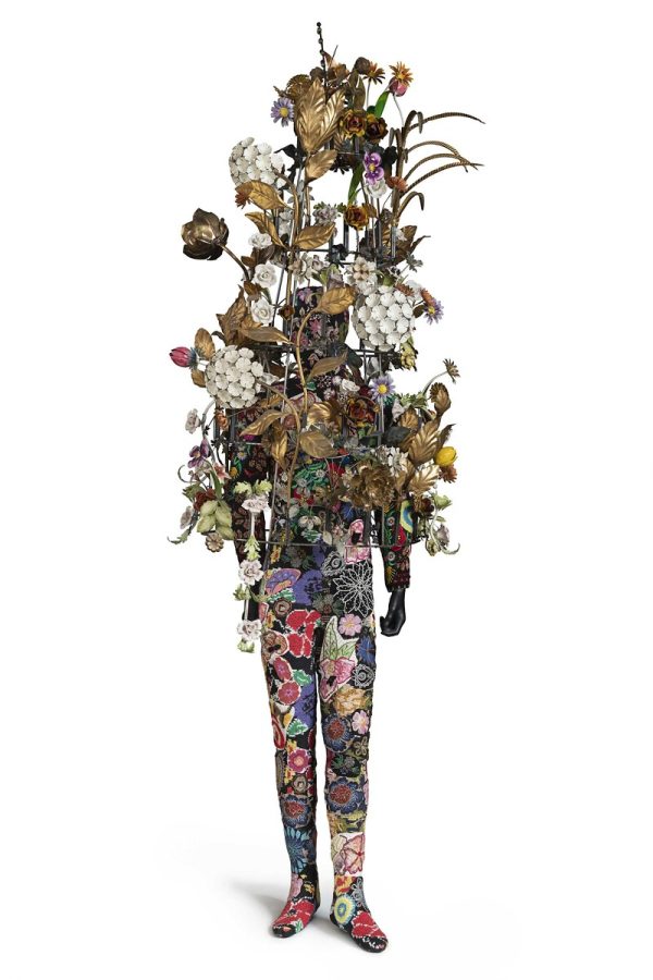 A mannequin's form dressed in a jumpsuit patterned in flowers. The top half is obscured by an large armature of white and gold flowers.