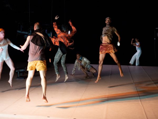 A group of barefoot dancers in athletic clothing on a dimly lit stage. Most of them have been captured mid-jump.