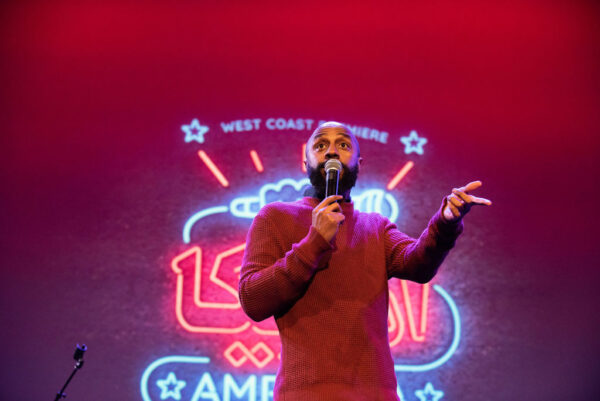 A male presenting person with light brown skin and a beard gestures while speaking into a microphone on a red lit stage in front of a large neon sign
