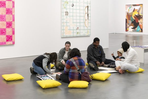 Adults and children sit on yellow pillows on a concrete floor doing art projects.