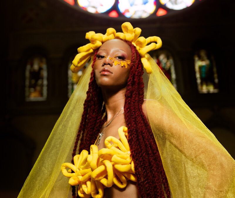 A female presenting person with light brown skin dressed in yellow with a yellow veil below a rosette style window.