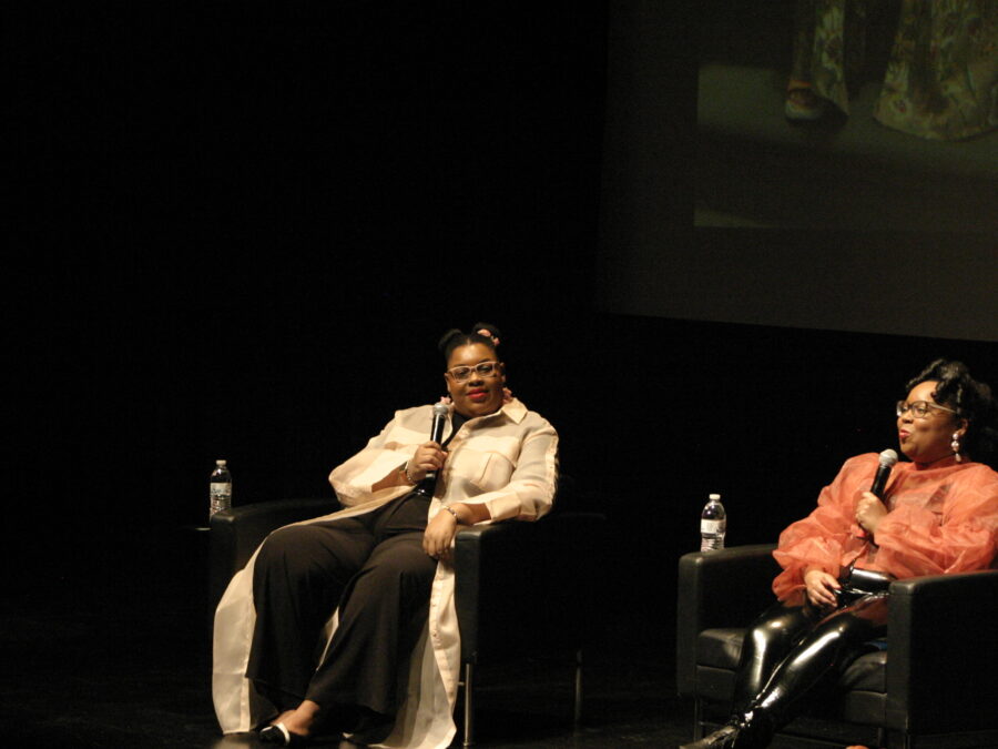 Two brown-skinned female presenting people sit on a darkened stage holding microphones