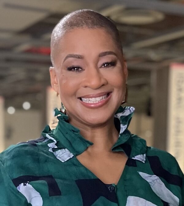 Portrait of a female-presenting person with a closely shaved head smiling widely. They wear a green collared shirt patterned with blue shapes.