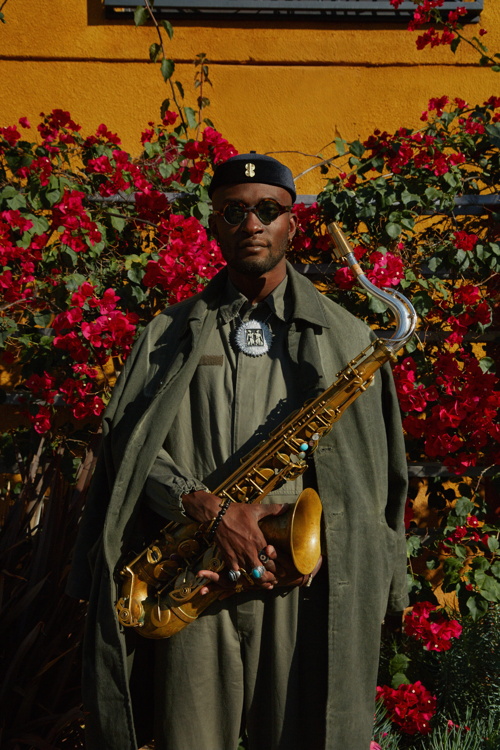 A dark brown-skinned man poses with a saxophone in front of a wall filled with flowers