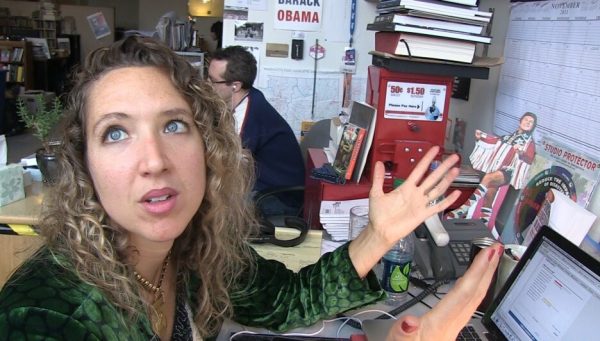 A female-presenting person gestures with their hands while looking up in a newsroom.