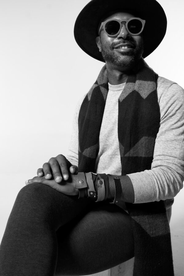 A black and white seated portrait of a brown-skinned male-presenting person wearing a wide brimmed hat and glasses