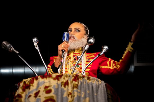 A female-presenting person with a fake grey beard and British red coat attire in front of a podium filled with microphones
