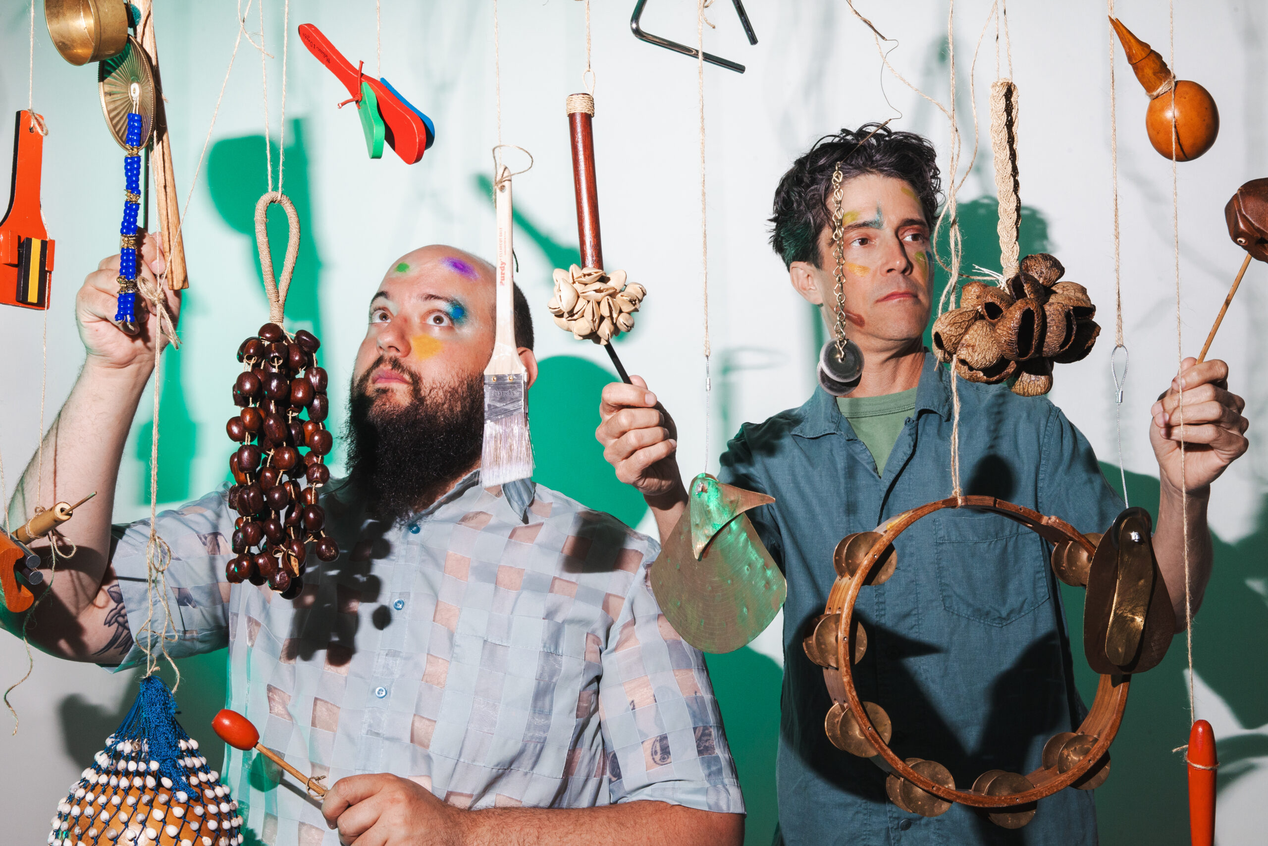 Two male-presenting people stand behind an array of various musical instruments hanging from strings