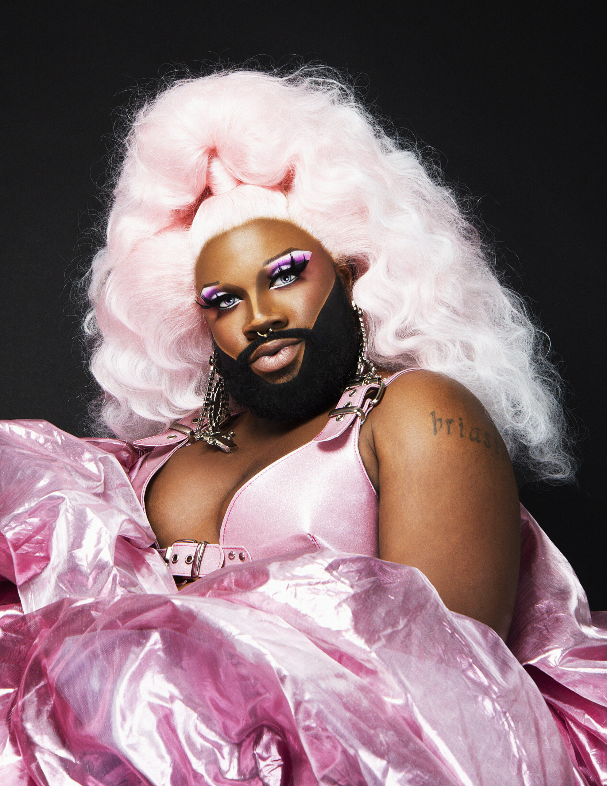 Three-quarter stylized portrait of a person with brown skin and a beard wearing a poofy pink evening gown with a voluminous, stylized pink hairdo, dramatic makeup, and accessorized with large earrings, posing against a black background.