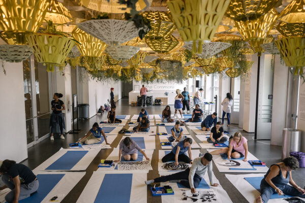 A group of people sit on yoga mats creating artwork in a room filled with planters hanging from the ceiling