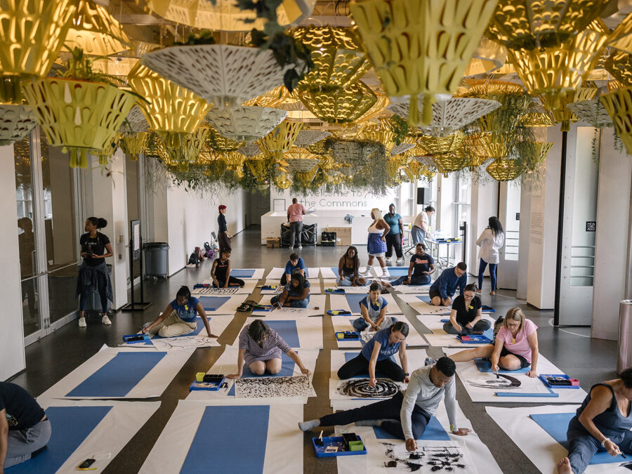 A group of people sit on yoga mats creating artwork in a room filled with planters hanging from the ceiling