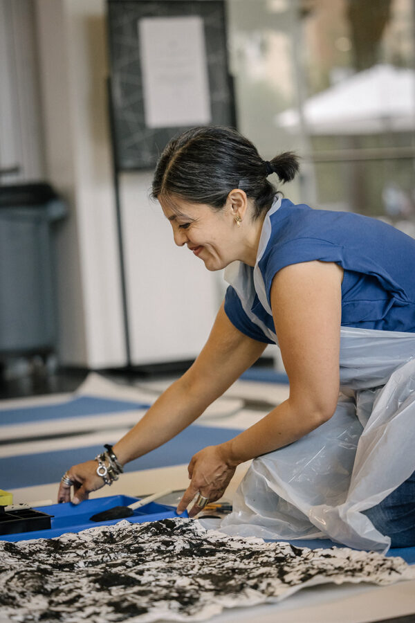 A female presenting person with olive skin smiles while adjusting a paint tray next to a black and white abstract artwork