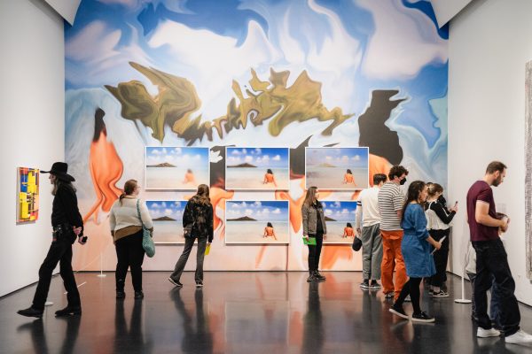 A group of people look at art in a room with a back wall covered in a colorful mural and pictures of a beach.