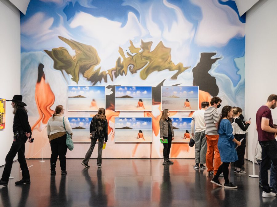 A group of people look at art in a room with a back wall covered in a colorful mural and pictures of a beach.