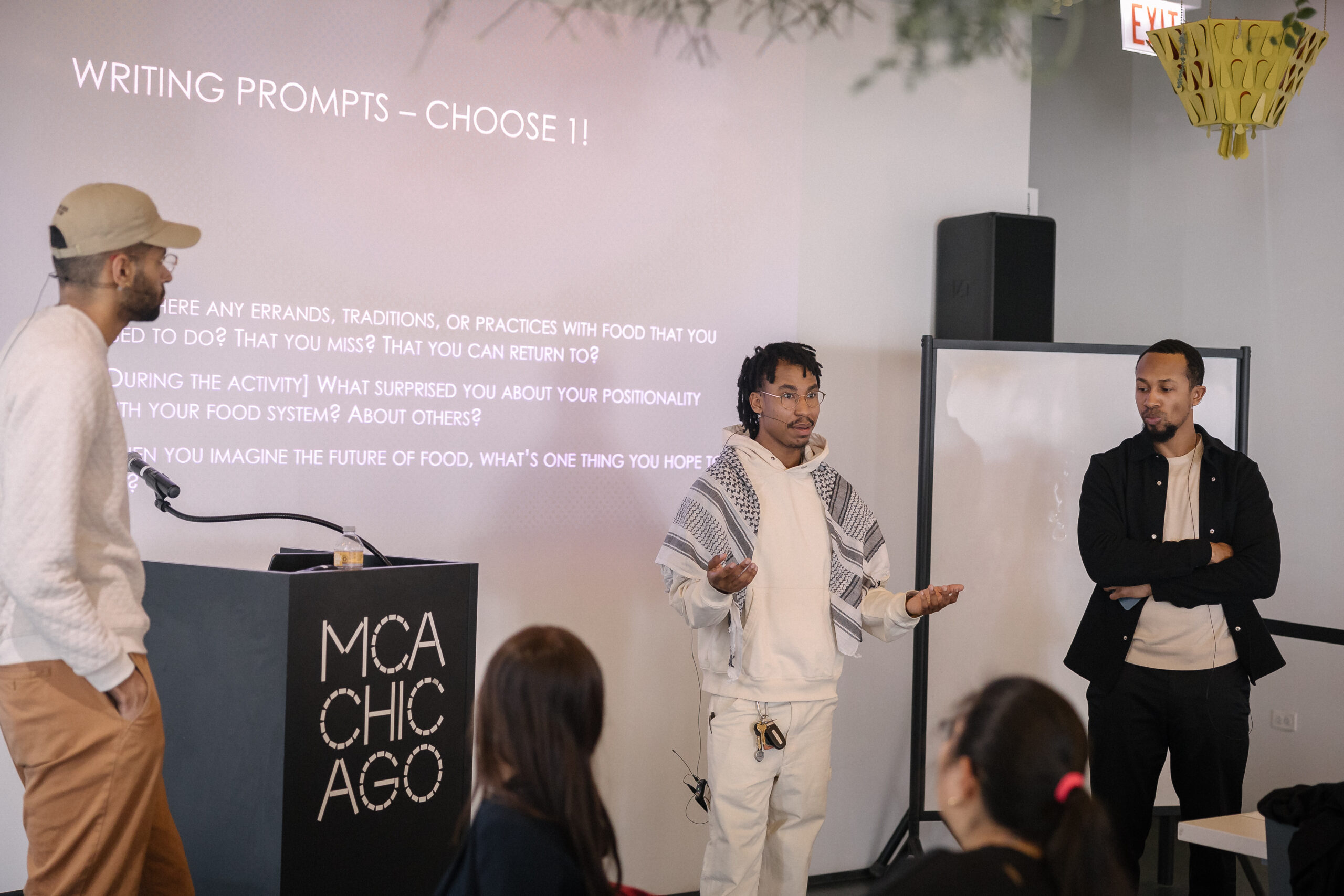 Three male-presenting people with light brown skin stand before an audience with a large screen on the wall behind them with a slide header of Writing Prompts