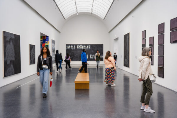 People in a bright, vaulted gallery with many dark works hung on three adjacent walls