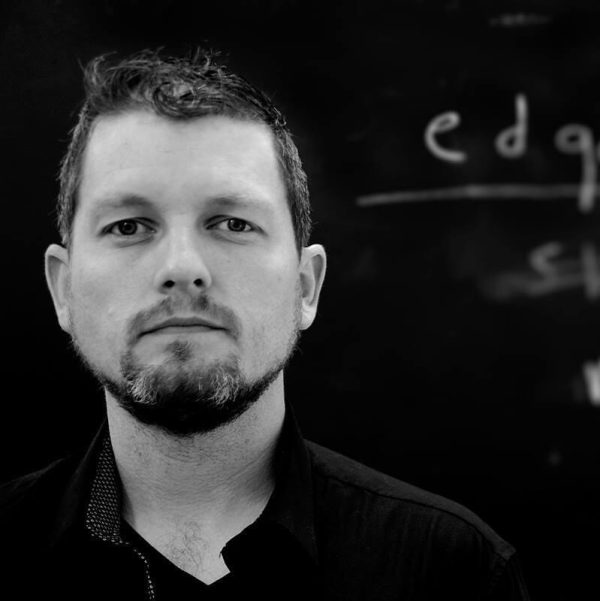 Black and white portrait of a light-skinned man with a goatee near a chalkboard with writing on it.